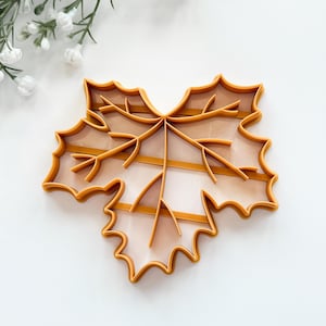 May include: An orange maple leaf cookie cutter. The cutter is made of plastic and has a detailed design of the leaf's veins. The cutter is on a white surface with white flowers and green stems in the background. Perfect for fall baking.