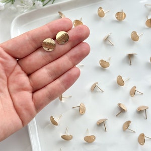 May include: Gold-colored earring studs with a textured, circular design. The earrings are displayed on a white tray, with several other studs scattered around. The studs have a post for insertion into the ear.