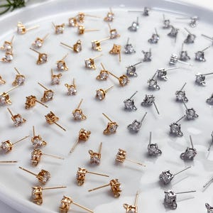 May include: A collection of gold-toned and silver-toned stud earring findings, each featuring a single clear round cubic zirconia stone set in a four-prong basket with a small loop at the base for attaching additional jewellery components.