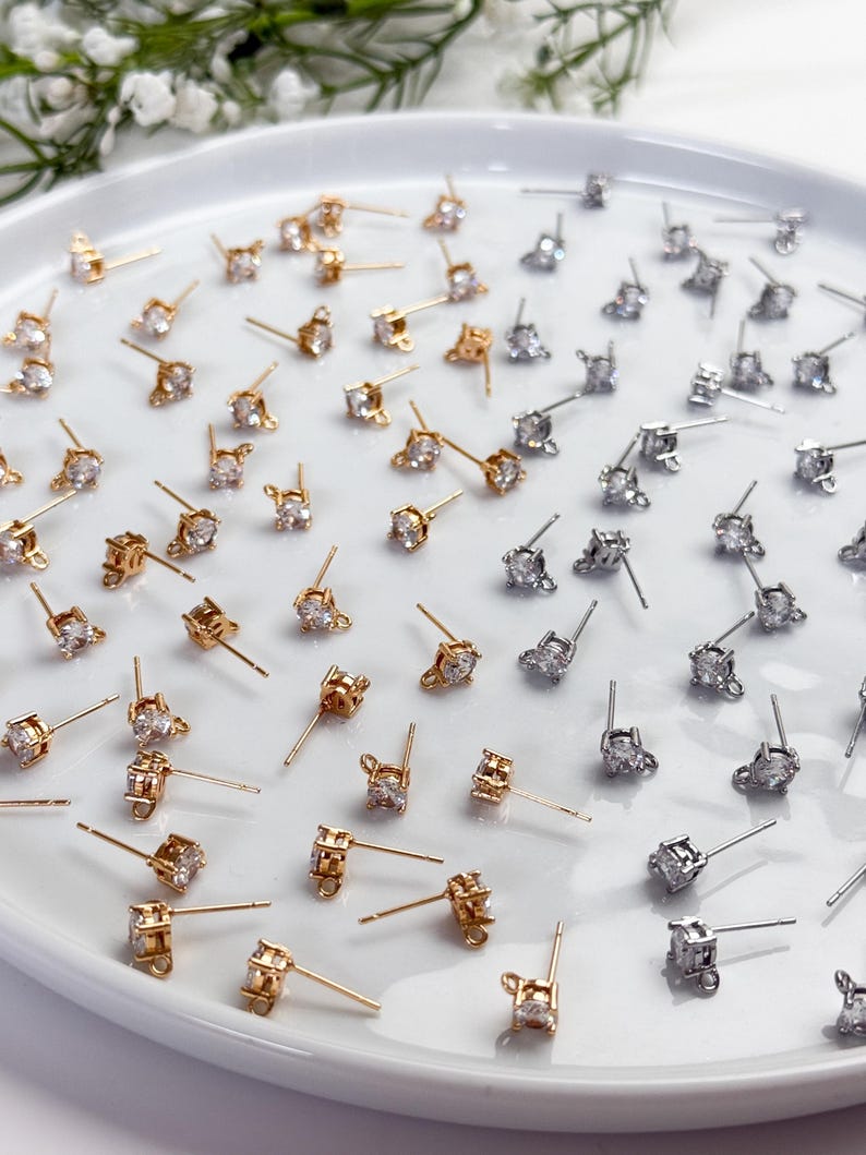 May include: A collection of gold-toned and silver-toned stud earring findings, each featuring a single clear round cubic zirconia stone set in a four-prong basket with a small loop at the base for attaching additional jewellery components.