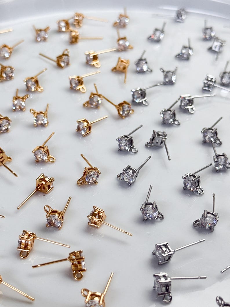 May include: A collection of stud earrings featuring clear, round-cut cubic zirconia stones set in metal prongs. The assortment includes both gold-toned and silver-toned metal finishes, each with a small loop at the base for attaching additional charms.