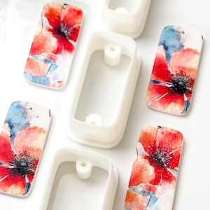 Puede incluir: Moldes rectangulares de plástico blanco e insertos a juego con estampado floral. Los insertos presentan un diseño de acuarela de amapolas rojas con centros negros y acentos azules y naranjas. Los moldes están vacíos y tienen forma rectangular.