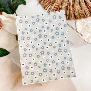Könnte beinhalten: Ein Blatt aus transparentem Vinyl mit einem sich wiederholenden Muster aus kleinen blauen und weißen Blumen auf hellgrauem Hintergrund.