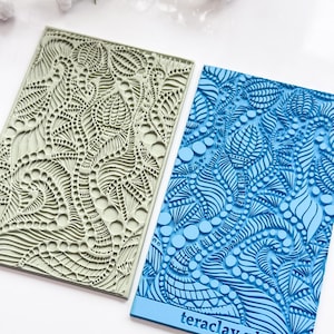 May include: Two silicone embossing mats for crafting. The mat on the left is light green with a swirling pattern of dots and lines. The mat on the right is blue with a similar pattern. The blue mat has the text "teraclay.nl" printed on the bottom.