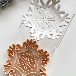 May include: Two snowflake-shaped cookie cutters. One is white with black speckles and rests on a clear acrylic stand. The other is a light peach colour. Both cutters have a detailed snowflake design with triangular and curved elements.