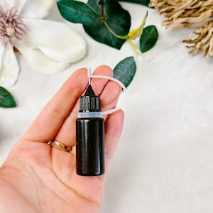 Könnte beinhalten: Eine kleine schwarze Flasche mit Tropfer und einem weißen Kunststoff-Schlaufen-Griff. Die Flasche wird in der Hand einer Person gehalten.