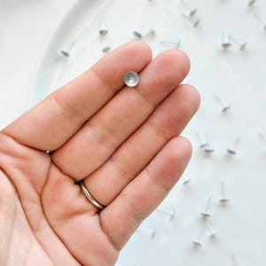 May include: Close-up of small, silver earring posts with flat, circular tops. One is held in a hand, others scattered on a white surface. These minimalist posts are ideal for crafting earrings. The posts are made of a silver-colored metal.
