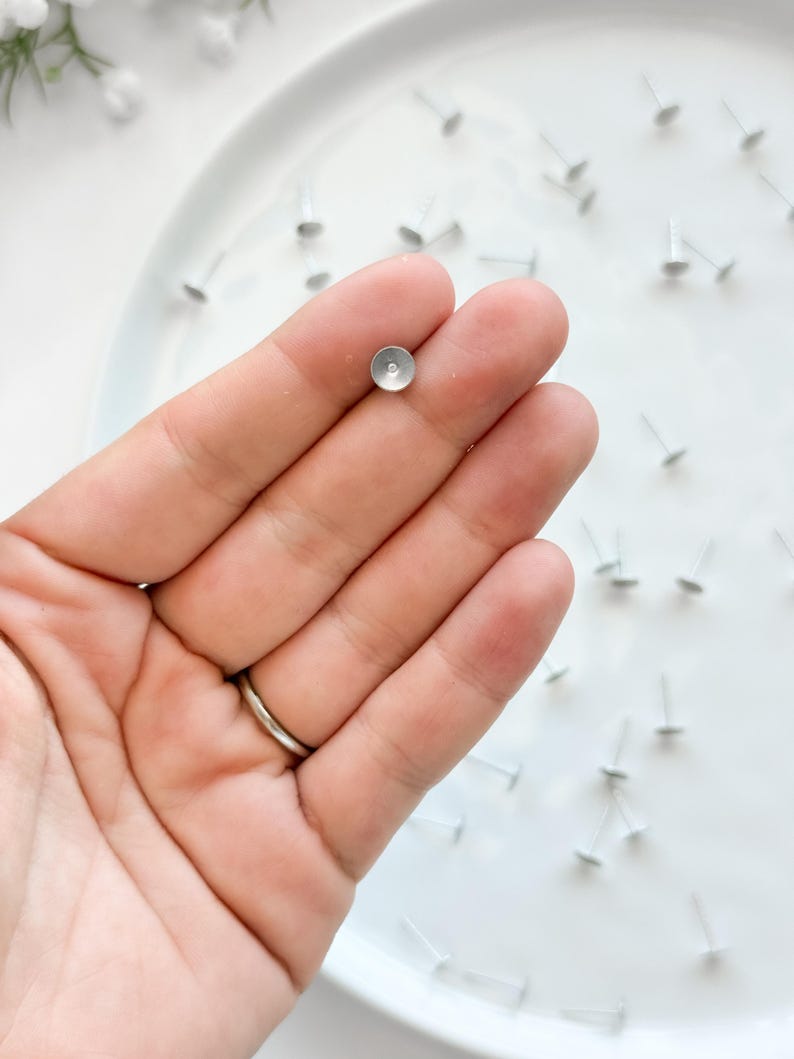 May include: Close-up of small, silver earring posts with flat, circular tops. One is held in a hand, others scattered on a white surface. These minimalist posts are ideal for crafting earrings. The posts are made of a silver-colored metal.