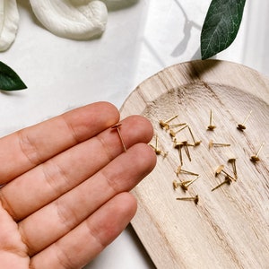 May include: A close-up of a hand holding a single gold earring post with a flat disc head. A wooden tray with a scattering of similar earring posts is in the background.