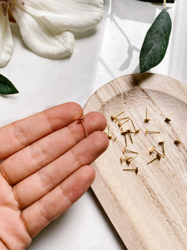 May include: A close-up of a hand holding a single gold earring post with a flat disc head. A wooden tray with a scattering of similar earring posts is in the background.