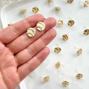 May include: A close-up of a hand holding two gold-toned, textured, round earring blanks. The blanks have a small hole in the center for attaching earring posts. The blanks are surrounded by other earring blanks on a white surface.