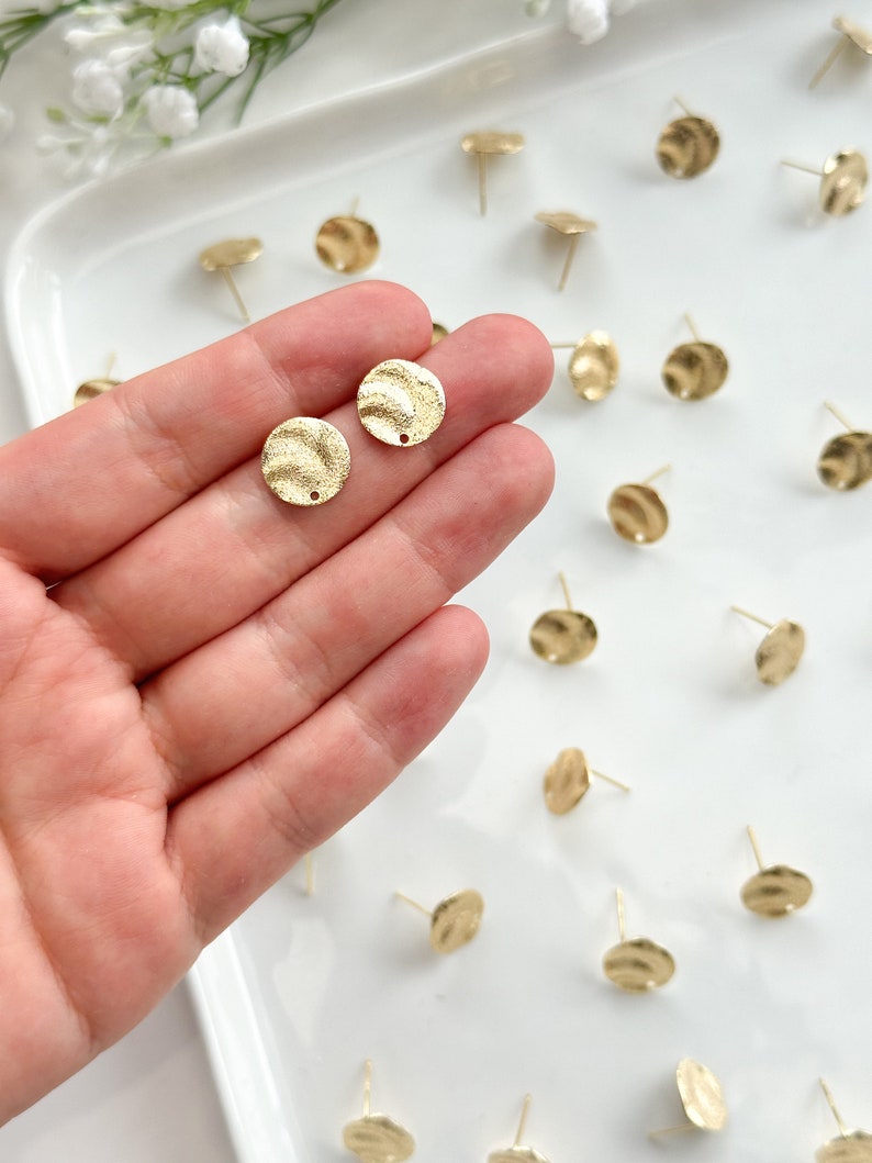 May include: A close-up of a hand holding two gold-toned, textured, round earring blanks. The blanks have a small hole in the center for attaching earring posts. The blanks are surrounded by other earring blanks on a white surface.