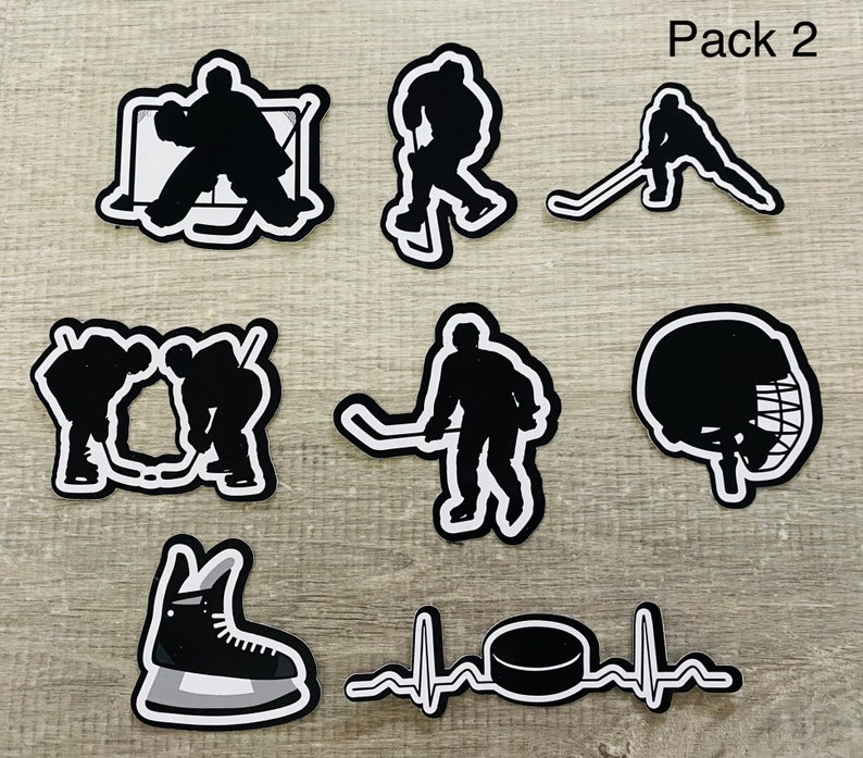 Pu&ograve; includere: Un set di nove adesivi a tema hockey in bianco e nero. Gli adesivi includono un portiere davanti a una rete, un giocatore che pattina con un disco, un giocatore che tira il disco, due giocatori che si passano il disco, un casco da hockey, un pattino da hockey e un disco da hockey con una grafica di battito cardiaco. Il testo "Pack 2" &egrave; visibile nell'angolo in alto a destra dell'immagine.