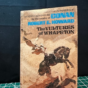 Könnte beinhalten: Ein Vintage-Paperback-Buchcover mit einer Fantasy-Abenteuergeschichte mit dem Titel "The Vultures of Whapeton" von Robert E. Howard. Das Cover zeigt eine skelettartige Figur, die ein schwarzes Pferd reitet, mit einem großen Vogel, der darüber fliegt.