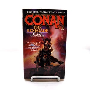 May include: A paperback book titled "CONAN THE RENEGADE" by Leonard Carpenter. The cover features a warrior on horseback with a bow and arrow, set against a fiery background. The book is displayed on a white stand.