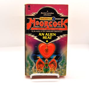 May include: A paperback book cover with a purple, red, and black background. The title is "An Alien Heat" and the author is Michael Moorcock. The cover features a red heart with a person inside and a woman with wings.