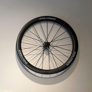 以下が含まれることがあります： リムに白い文字で「POLYMER」と書かれた黒い自転車のホイール。ホイールには、中央のハブから放射状に伸びる細い黒いスポークがあります。ホイールは白い壁に取り付けられています。