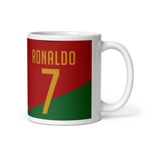 K&ouml;nnte beinhalten: Wei&szlig;e Keramiktasse mit rotem und gr&uuml;nem Design. Die Tasse zeigt den Namen "RONALDO" und die Nummer "7" in Gelb.