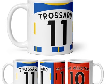 May include: Three white ceramic mugs with different designs. The first mug has a blue and yellow design with the text "TROSSARD 11". The second mug has a blue and yellow design with the text "TROSSARD 11". The third mug has a red design with the text "MAC ALLISTER 10".