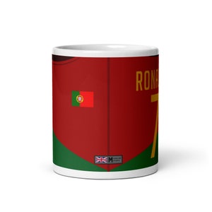 K&ouml;nnte beinhalten: Wei&szlig;e Keramiktasse mit einem rot-gr&uuml;n-goldenen Design. Die Tasse zeigt ein Fu&szlig;balltrikot mit der Nummer 7 und dem Namen "RONALDO" darauf gedruckt. Das Trikot hat auch die portugiesische Flagge auf der linken Seite. Die Tasse hat ein kleines Etikett auf der Unterseite, auf dem "SIDELINE SPORTS" steht.