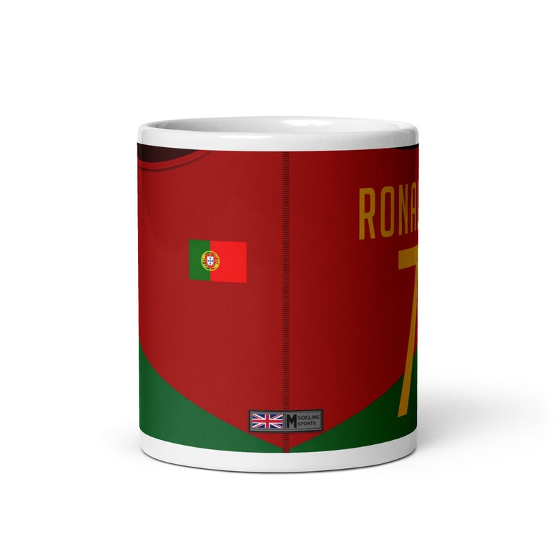 K&ouml;nnte beinhalten: Wei&szlig;e Keramiktasse mit einem rot-gr&uuml;n-goldenen Design. Die Tasse zeigt ein Fu&szlig;balltrikot mit der Nummer 7 und dem Namen "RONALDO" darauf gedruckt. Das Trikot hat auch die portugiesische Flagge auf der linken Seite. Die Tasse hat ein kleines Etikett auf der Unterseite, auf dem "SIDELINE SPORTS" steht.