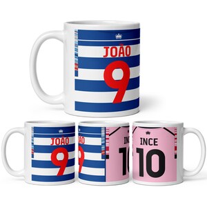 May include: Four white ceramic mugs with different designs. The first mug has a blue and white striped design with the text "JOÃO 9" in red. The second mug has a similar design with the text "JOÃO 9". The third mug has a pink design with the text "INCE 10" in black. The fourth mug has a similar design with the text "INCE 10".
