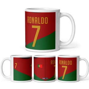 K&ouml;nnte beinhalten: Eine wei&szlig;e Keramiktasse mit einem roten, gr&uuml;nen und goldenen Design. Die Tasse tr&auml;gt den Namen "RONALDO" und die Nummer "7" in goldenen Buchstaben. Die Tasse ist ein tolles Geschenk f&uuml;r Fu&szlig;ballfans.