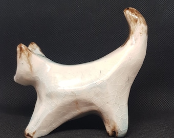 May include: A white ceramic cat figurine with brown accents. The cat is standing on all four legs with its tail curled up. The figurine has a crackled glaze finish.