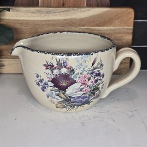 May include: Cream-colored ceramic gravy boat with a floral design featuring purple, pink, and blue flowers. The rim has a dark blue, dotted pattern. It has a spout and a handle, perfect for serving sauces and gravies.