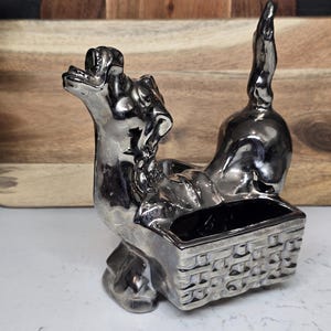 May include: A silver-colored ceramic dog figurine with a basket. The dog is in a playful pose, with its tail raised. The basket is rectangular with a woven design. The figurine is a decorative item.