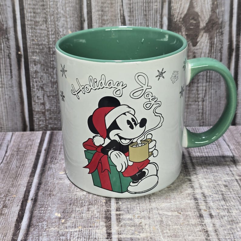 K&ouml;nnte beinhalten: Wei&szlig;er Keramikbecher mit gr&uuml;nem Innenraum und Henkel. Der Becher zeigt eine Grafik von Mickey Mouse mit Weihnachtsm&uuml;tze, die einen Becher hei&szlig;e Schokolade h&auml;lt, mit den Worten "Holiday Joy" und Schneeflocken. Der Becher steht auf einer Holzoberfl&auml;che.