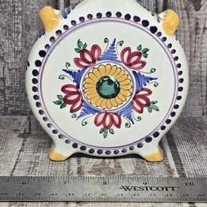 May include: A round ceramic flask with a cork stopper, featuring a hand-painted floral pattern in yellow, red, blue, and green on a white background. The flask has yellow handles and feet, and a metal ruler shows it is approximately 8 inches wide.