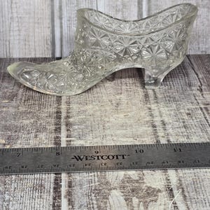 May include: A clear glass shoe figurine featuring a textured starburst pattern across its surface. The shoe is positioned above a metal Westcott ruler, which indicates the object measures approximately 6 inches in total length from toe to heel.
