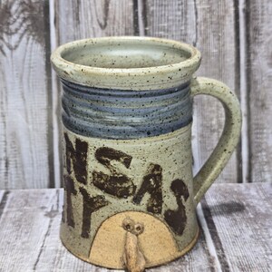 May include: A handmade ceramic mug with a beige and blue color scheme. The mug features the words "Kansas City" in brown lettering and a decorative owl design. The mug has a handle and is suitable for hot or cold beverages.