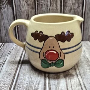 May include: A ceramic gravy boat with a reindeer design. The reindeer has brown antlers, a brown face, a red nose, and a green bow tie. The gravy boat is cream colored with blue stripes.