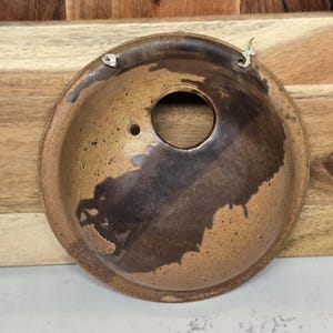 May include: A handmade, ceramic, brown-toned bowl with a large central hole and a smaller hole. The bowl has a dark brown, abstract design on the interior. Two small metal pieces are attached to the rim for hanging.