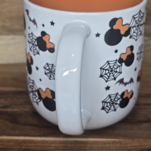May include: White ceramic mug with an orange interior. The exterior features black Mickey Mouse silhouettes with orange bows, spiderwebs, bats, and stars. The mug has a white handle and is set against a wooden surface.