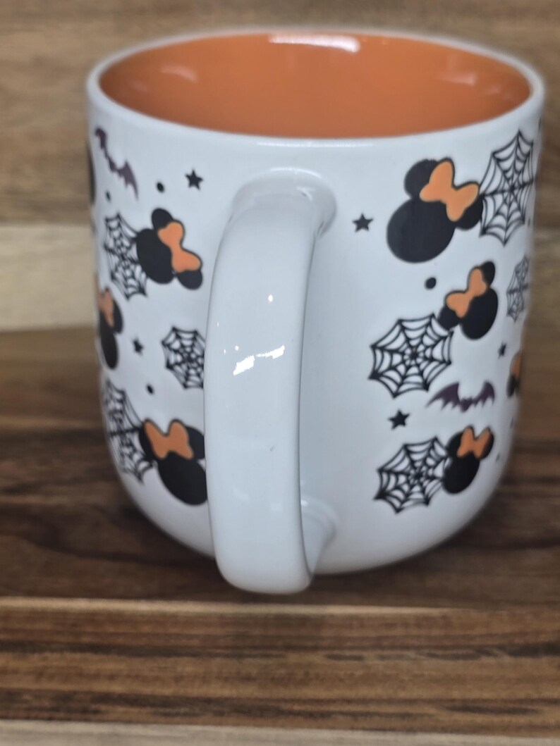 May include: White ceramic mug with an orange interior. The exterior features black Mickey Mouse silhouettes with orange bows, spiderwebs, bats, and stars. The mug has a white handle and is set against a wooden surface.