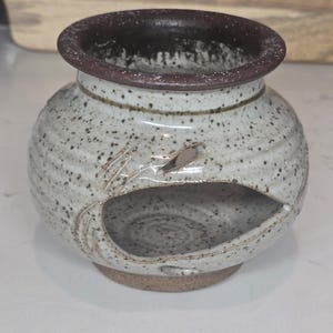 May include: A speckled ceramic oil warmer with a dark brown rim and a leaf-shaped opening. The small, round warmer has a circular base and is designed for use with essential oils. The exterior is a speckled white and gray.