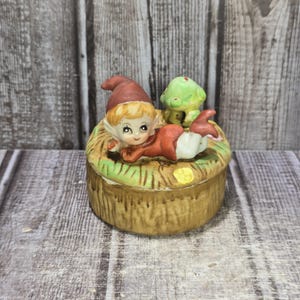 May include: A ceramic trinket box featuring a whimsical elf figure in a red outfit and hat, lying on a mushroom-shaped base. The box is painted with earthy tones of brown, green, and yellow, with a green mushroom beside the elf.
