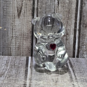 May include: A clear glass bear figurine with a small red heart on its chest. The bear is sitting upright with its arms and legs visible. The background is a wooden surface.