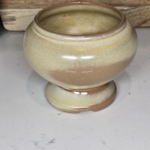 May include: A small, round, ceramic planter with a brown and beige glaze. The planter has a wide base and a narrow opening.