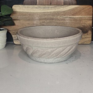 May include: A light beige ceramic bowl with a textured, twisted rope design around the base. The bowl has a wide opening and is set against a wooden cutting board and a black tiled backsplash.