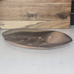 May include: A brown and tan ceramic leaf-shaped dish. The dish has a smooth, glossy finish and a shallow, elongated design. The dish is approximately 12 inches long and 5 inches wide. The background includes a wooden cutting board.