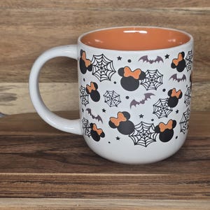 May include: White ceramic mug with a Halloween theme. The mug features a pattern of black Minnie Mouse heads with orange bows, spiderwebs, bats, and stars. The interior is orange.