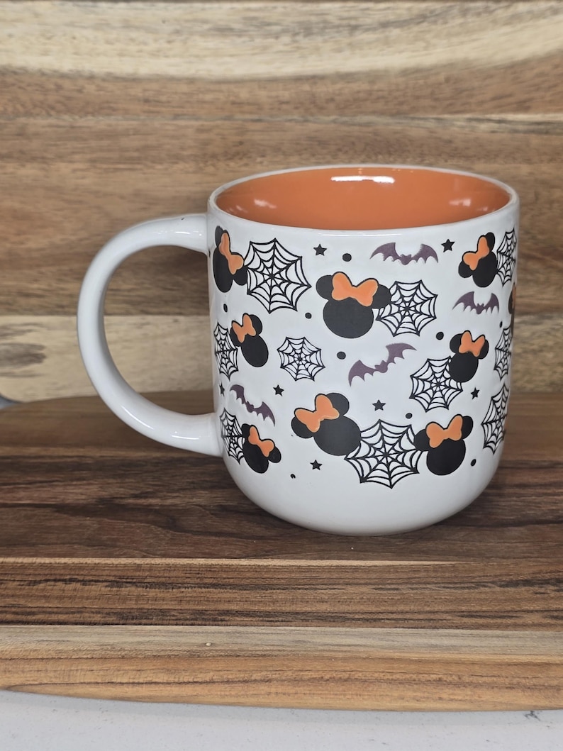 May include: White ceramic mug with a Halloween theme. The mug features a pattern of black Minnie Mouse heads with orange bows, spiderwebs, bats, and stars. The interior is orange.