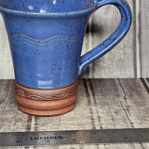 May include: A blue ceramic mug with dark speckles, featuring a wavy embossed line near the rim and a textured, unglazed terracotta-colored base with a decorative band. A metal ruler is positioned in front, showing a width of about 4 inches.