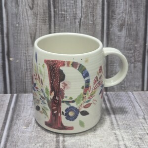 May include: White ceramic mug with a large letter "P" in a floral design. The letter is red with a colorful striped border. The mug has a handle and is decorated with flowers and leaves in various colors.