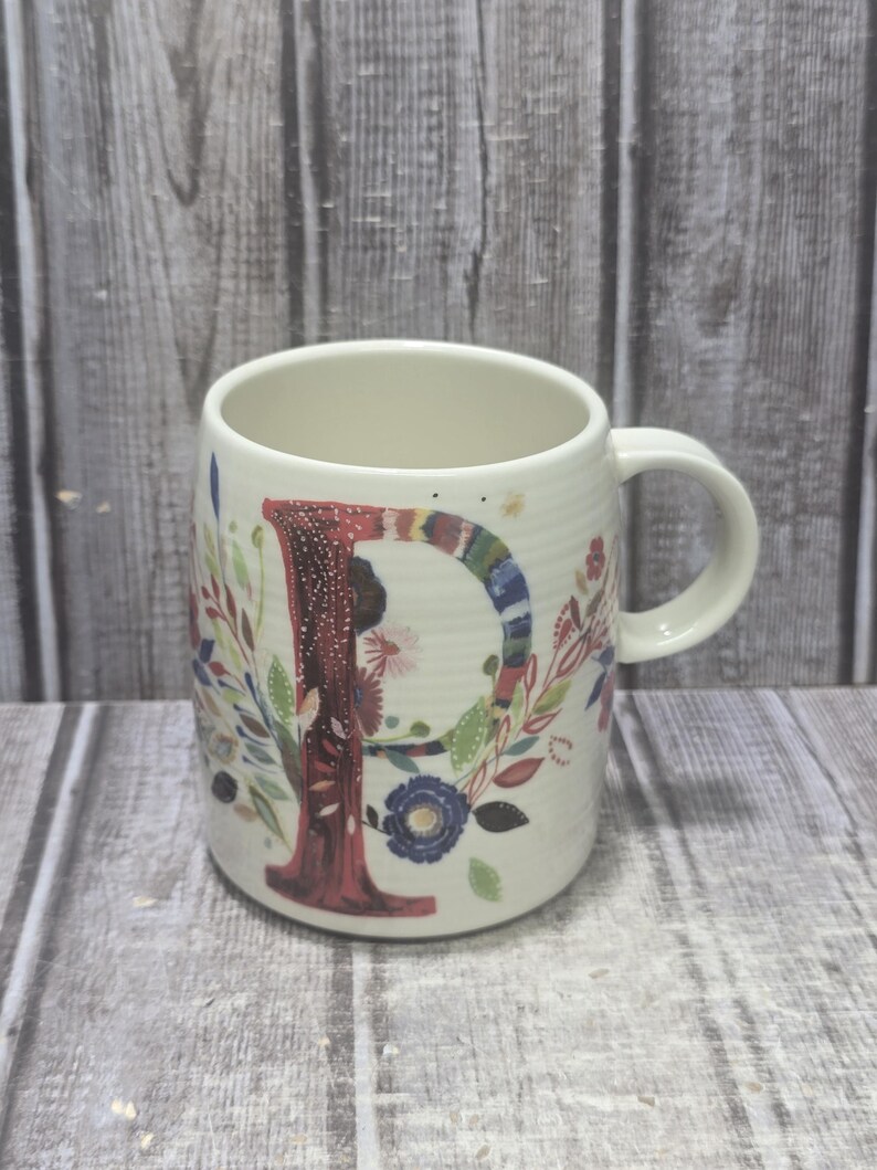 May include: White ceramic mug with a large letter "P" in a floral design. The letter is red with a colorful striped border. The mug has a handle and is decorated with flowers and leaves in various colors.