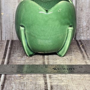 May include: A glossy green ceramic planter with a scalloped rim and three short legs. A metal Westcott ruler is positioned in front, showing the object spans approximately 4 inches in width from the 6-inch to the 10-inch mark on the scale.